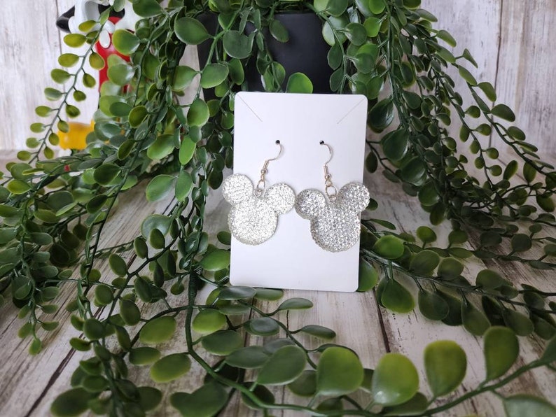 May include: Silver glitter Mickey Mouse earrings on a white card. The earrings are hanging from gold hooks.