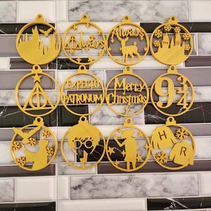 May include: A collection of gold-colored, circular Christmas ornaments with various designs and text. Designs include a castle, a stag, the words "Always" and "Merry Christmas", and the number "9 3/4". Other ornaments feature a lightning bolt, a silhouette of a person, and the letters "H" and "R".