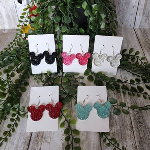 May include: Six pairs of dangle earrings featuring a Mickey Mouse head design. The earrings are made of glitter and come in black, silver, red, turquoise, and pink.