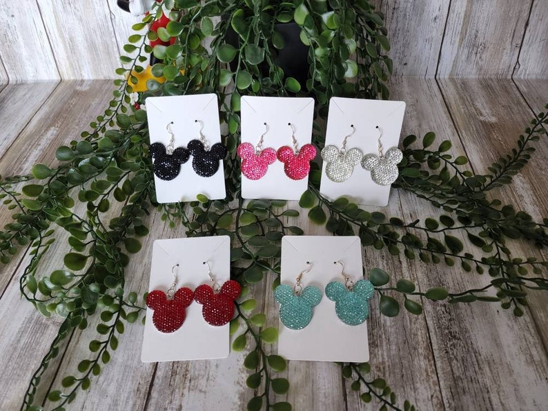 May include: Six pairs of dangle earrings featuring a Mickey Mouse head design. The earrings are made of glitter and come in black, silver, red, turquoise, and pink.