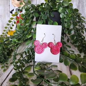 May include: Pink sparkly Mickey Mouse earrings on a white card with a green plant in the background.