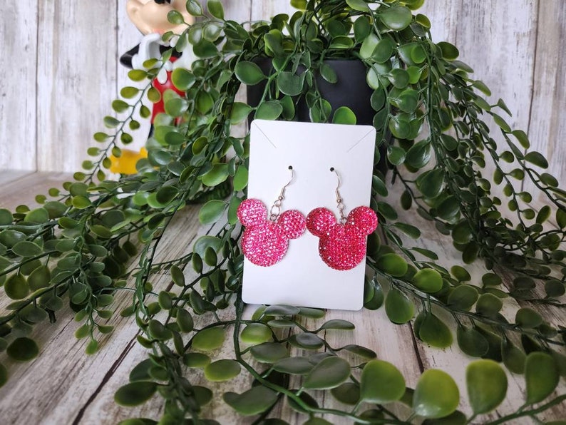 May include: Pink sparkly Mickey Mouse earrings on a white card with a green plant in the background.