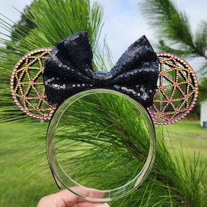 May include: Black sequined bow headband with pink and yellow geometric ears. The headband is on a clear display stand.