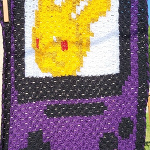 May include: Crocheted blanket with a purple, black, and white design. The blanket features a pixelated image of a yellow character with red cheeks, resembling a video game character.