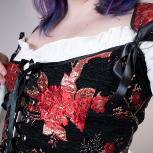 May include: A black corset with red and gold floral embroidery and black satin ribbon ties. The corset has a lace-up front and is worn over a white blouse.