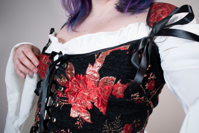 May include: A black corset with red and gold floral embroidery and black satin ribbon ties. The corset has a lace-up front and is worn over a white blouse.