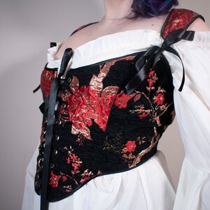 May include: A black corset with red and gold floral embroidery and black ribbon ties. The corset is worn over a white blouse with puffed sleeves.