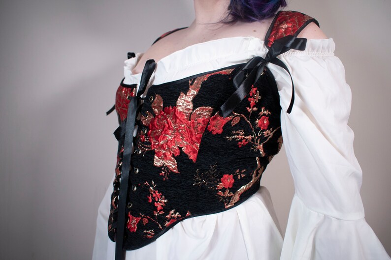 May include: A black corset with red and gold floral embroidery and black ribbon ties. The corset is worn over a white blouse with puffed sleeves.