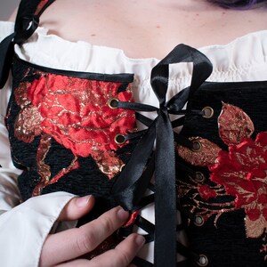 May include: A black corset with red and gold floral embroidery and black ribbon lacing. The corset has a white ruffle trim at the top.