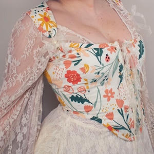 May include: A white corset with a floral print featuring red, orange, yellow, and green flowers. The corset has a lace-up front and is worn with a white lace top and skirt.