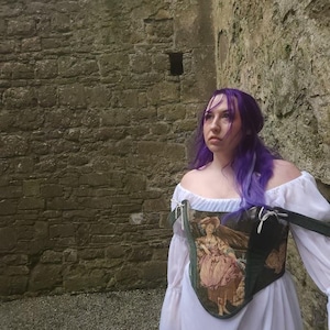 May include: A woman with long purple hair wearing a white off-the-shoulder dress with a tapestry corset featuring a floral design and a woman in a dress.