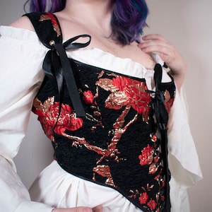 May include: A black corset with red and gold floral embroidery. The corset has a lace-up front and is worn over a white blouse.