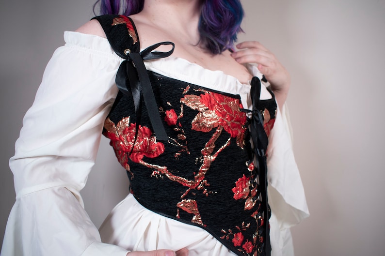 May include: A black corset with red and gold floral embroidery. The corset has a lace-up front and is worn over a white blouse.