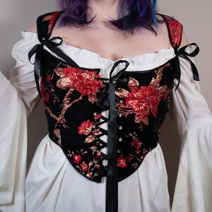 May include: A black corset with red and gold floral embroidery and black ribbon lacing. The corset has a sweetheart neckline and is worn over a white blouse with ruffled sleeves.
