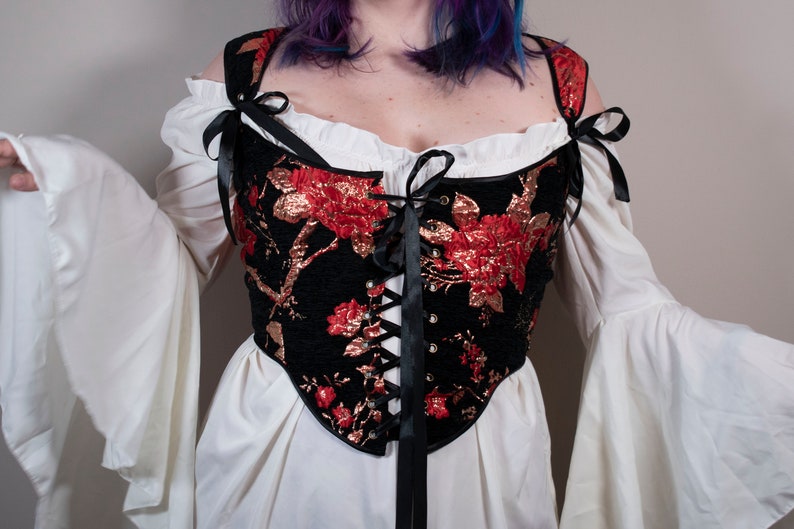 May include: A black corset with red and gold floral embroidery and black ribbon lacing. The corset has a sweetheart neckline and is worn over a white blouse with ruffled sleeves.
