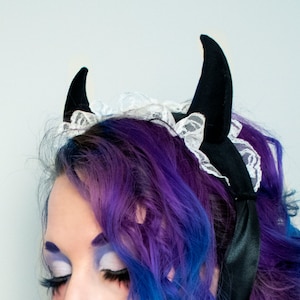 May include: A woman with purple and blue hair is wearing a black headband with devil horns and a white lace trim.