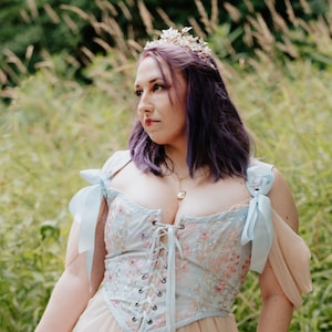 May include: A woman with purple hair wearing a light blue and pink floral corset with a light blue ribbon on each shoulder. She is wearing a crown and looking to the right.
