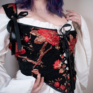 May include: A black corset with red and gold floral embroidery and black ribbon lacing. The corset has a sweetheart neckline and is worn over a white blouse.