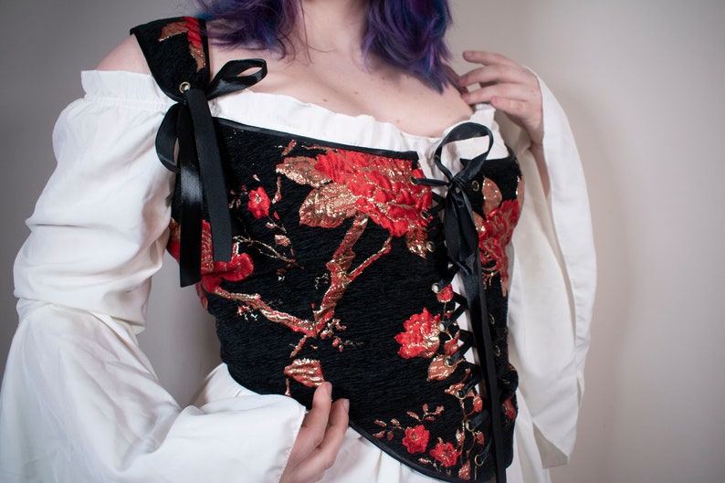 May include: A black corset with red and gold floral embroidery and black ribbon lacing. The corset has a sweetheart neckline and is worn over a white blouse.