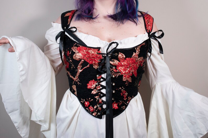 May include: A black corset with red and gold floral embroidery and black ribbon lacing. The corset is worn over a white blouse with large, ruffled sleeves.