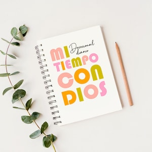 May include: A white spiral bound journal with the text "Devocional diario" and "MI TIEMPO CON DIOS" in colorful letters. The journal is on a white surface with a pencil.