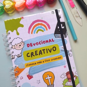 Könnte beinhalten: Ein Spiralnotizbuch mit den Worten "Devocional Creativo" und "Conoce más a Dios creando!" auf dem Einband. Der Einband zeigt einen Regenbogen, eine Schafillustration und andere farbenfrohe Grafiken. Ebenfalls sichtbar sind herzförmige Perlen und Stifte.
