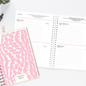 Puede incluir: Una agenda con espiral con una cubierta blanca con la palabra "BLESSED" en una fuente rosa ondulada. La agenda está abierta para mostrar páginas rayadas con fechas y notas. Se ve un bolígrafo y otros artículos de papelería.