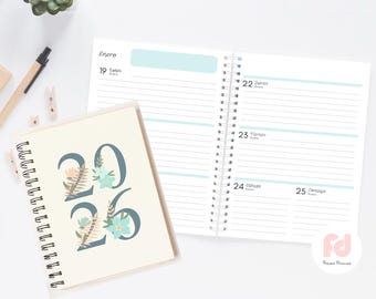 Pode incluir: Agenda de espiral creme com o ano "2026" em azul-petróleo, adornada com detalhes florais. A agenda aberta exibe páginas pautadas com cabeçalhos em azul claro. Uma caneta e clipes decorativos são visíveis.