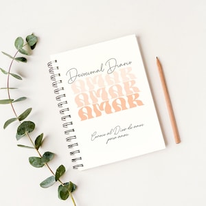 Op de afbeelding: Een wit spiraalnotitieblok met de tekst "Devocional Diario" en het woord "AMAR" dat vier keer wordt herhaald in een verloop van perzik en roze. De tekst "Conoce al Dios de amor para amar" staat in het zwart onderaan de pagina.