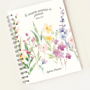 May include: A white spiral bound notebook with a floral design. The text on the cover reads "ÉL convierte desiertos en jardines. Isaiah 41:19" and "Agenda Perpetua".