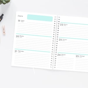 Pode incluir: Agenda de espiral aberta com p&aacute;ginas pautadas e detalhes em azul claro. A agenda est&aacute; aberta para uma vista semanal, com datas e dias da semana vis&iacute;veis. A palavra "Enero" est&aacute; no topo. Uma caneta e pequenos clipes de madeira est&atilde;o na superf&iacute;cie branca.