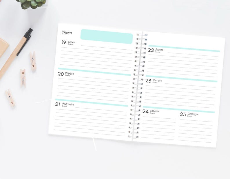 Pode incluir: Agenda de espiral aberta com p&aacute;ginas pautadas e detalhes em azul claro. A agenda est&aacute; aberta para uma vista semanal, com datas e dias da semana vis&iacute;veis. A palavra "Enero" est&aacute; no topo. Uma caneta e pequenos clipes de madeira est&atilde;o na superf&iacute;cie branca.