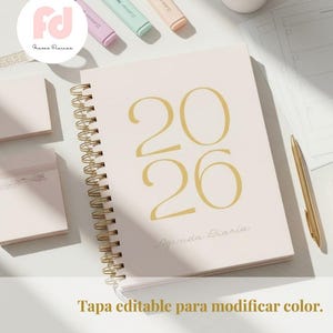 May include: A light pink spiral-bound planner with the gold numbers "2026" on the cover, along with the words "Agenda Diaria". The image also shows a gold pen, three highlighters, and a small plant.