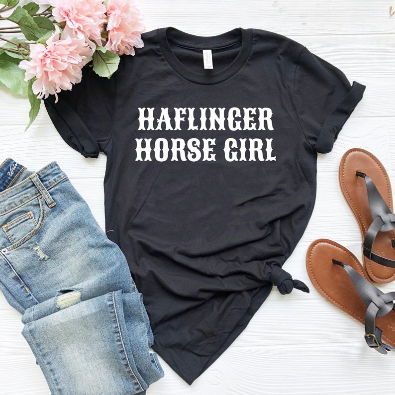 May include: Black t-shirt with white text that reads "HAFLINGER HORSE GIRL". The shirt is shown with blue jeans and brown sandals.