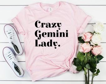 May include: A light pink t-shirt with the text "Crazy Gemini Lady" in black. The shirt is displayed with white sneakers and a bouquet of pink and white roses on a white wooden surface.