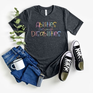 May include: A dark grey t-shirt with the text "Abilities Outweigh Disabilities" in colorful letters. The shirt is paired with blue jeans and black sneakers.