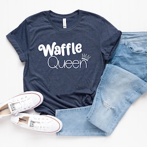 May include: A navy blue t-shirt with white text that reads "Waffle Queen" with a crown above the word "Queen". The shirt is paired with blue jeans and white sneakers.