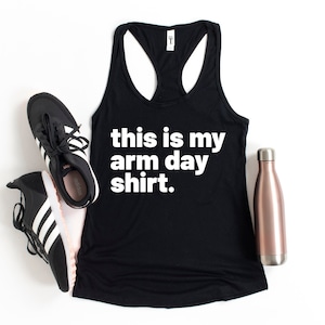 Puede incluir: Camiseta de tirantes negra con el texto "this is my arm day shirt." en ella. La camiseta está colocada sobre una superficie blanca con un par de zapatillas negras y una botella de agua de color oro rosa a su lado.