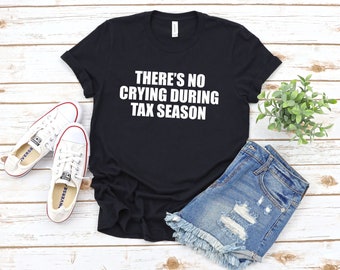 Könnte beinhalten: Schwarzes T-Shirt mit weißem Text, der "There's no crying during tax season" lautet. Das Shirt liegt flach auf einer Holzoberfläche mit einem Paar weißen Sneakers, Jeans-Shorts und einer kleinen Topfpflanze.