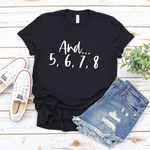 May include: Black t-shirt with white text that reads "And... 5, 6, 7, 8". The shirt is laying on a white wooden surface with a pair of white sneakers and denim shorts.