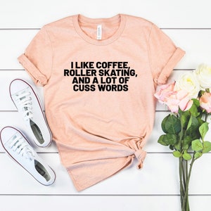 Op de afbeelding: Een perzikkleurig T-shirt met de tekst "Ik hou van koffie, rolschaatsen en veel scheldwoorden" in het zwart.