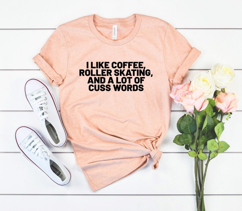 Op de afbeelding: Een perzikkleurig T-shirt met de tekst "Ik hou van koffie, rolschaatsen en veel scheldwoorden" in het zwart.