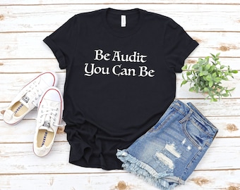 May include: Black t-shirt with white text that reads "Be Audit You Can Be". The shirt is laying on a wooden surface with a pair of white sneakers, denim shorts, and a potted plant.