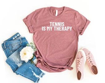 May include: A mauve t-shirt with the text "Tennis is my therapy" in white lettering. The shirt is laid flat on a white background with a pair of pink trainers and blue jeans.
