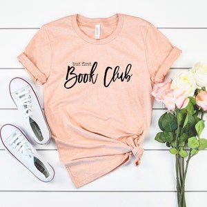 May include: A peach-coloured t-shirt with the text "but first, Book Club" in black lettering. The shirt is laid out on a white wooden surface with a pair of white trainers and a bouquet of pink and white roses.