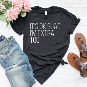 May include: Black t-shirt with white text that reads "It's ok guac I'm extra too". The shirt is laid out on a light wood surface with a pair of blue jeans and brown sandals.