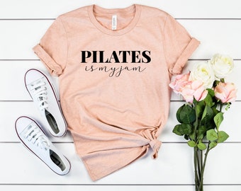 May include: A peach-colored t-shirt with the text "PILATES is my jam" in black lettering. The shirt is laid out on a white surface with a pair of white sneakers and a bouquet of pink and white flowers.