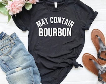 May include: Black t-shirt with white text that reads "May Contain Bourbon". The shirt is laid out on a white surface with a pair of blue jeans and sandals.