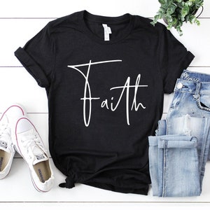 Pode incluir: Camiseta preta com um gr&aacute;fico branco em it&aacute;lico de "Faith". A camiseta est&aacute; sobre uma superf&iacute;cie branca com um par de t&ecirc;nis brancos e uma cal&ccedil;a jeans azul.