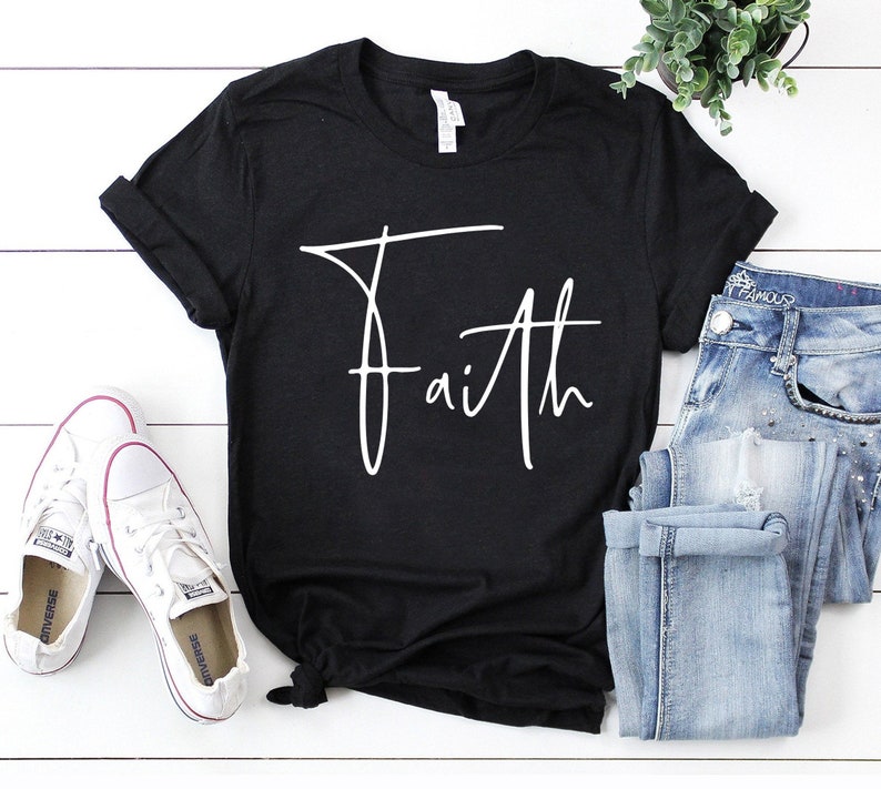 Pode incluir: Camiseta preta com um gr&aacute;fico branco em it&aacute;lico de "Faith". A camiseta est&aacute; sobre uma superf&iacute;cie branca com um par de t&ecirc;nis brancos e uma cal&ccedil;a jeans azul.